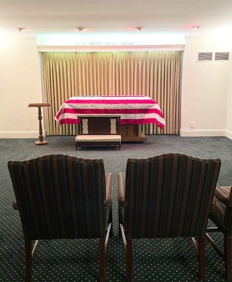A funeral viewing room with a flag-draped casket on a raised platform, two chairs in the foreground.