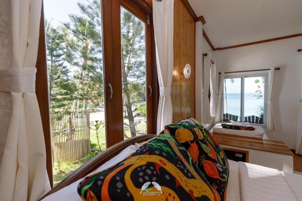 Bedroom with large windows, view of greenery and ocean, colorful pillow, wooden frame.