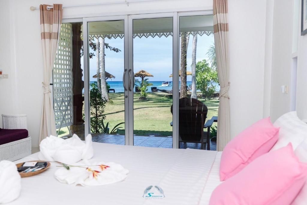 Bedroom with a view of the ocean. White bed, pink pillows, sliding glass doors, and a lush green yard.