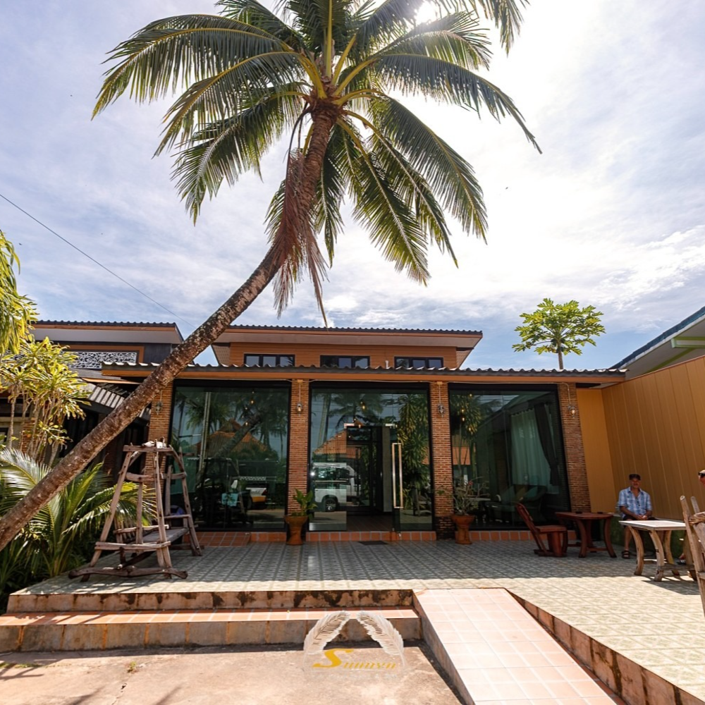 Palm tree leans over a building with glass doors, outdoor seating, and a man seated at a table.