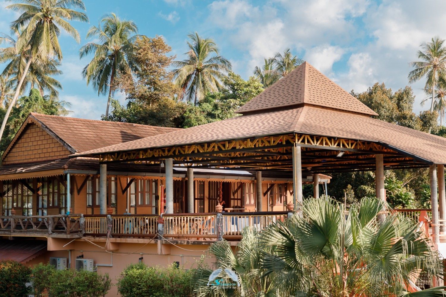Tropical building with brown tiled roof, wooden deck, and palm trees under a blue sky.