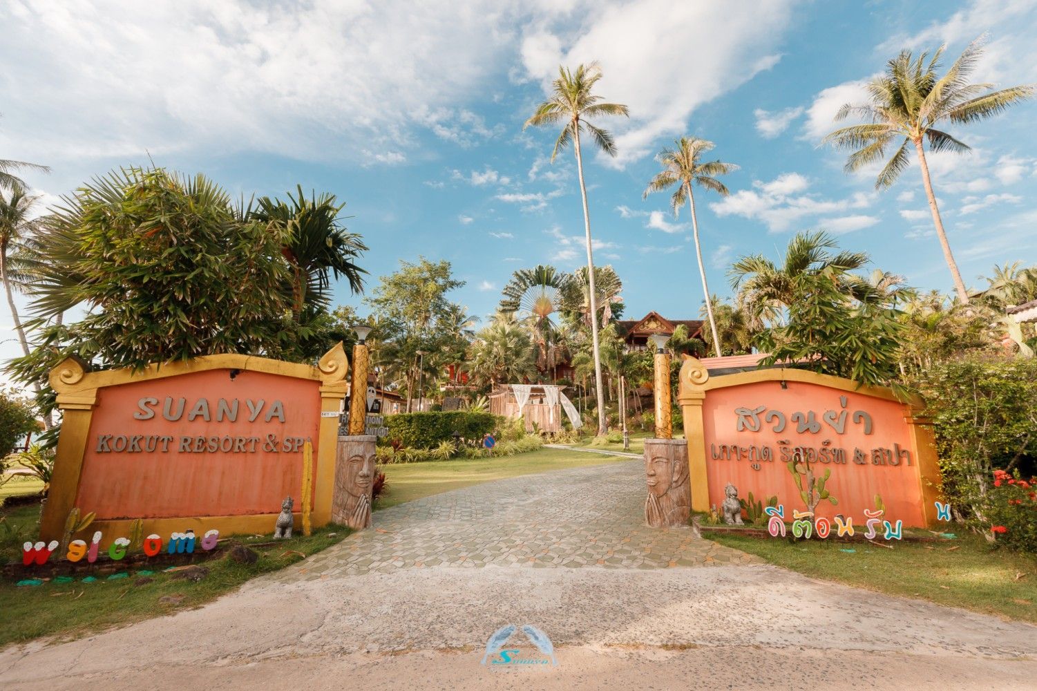 Entrance to "Suanya Resort & Beach Club" with ornate signs, palm trees, and blue sky.