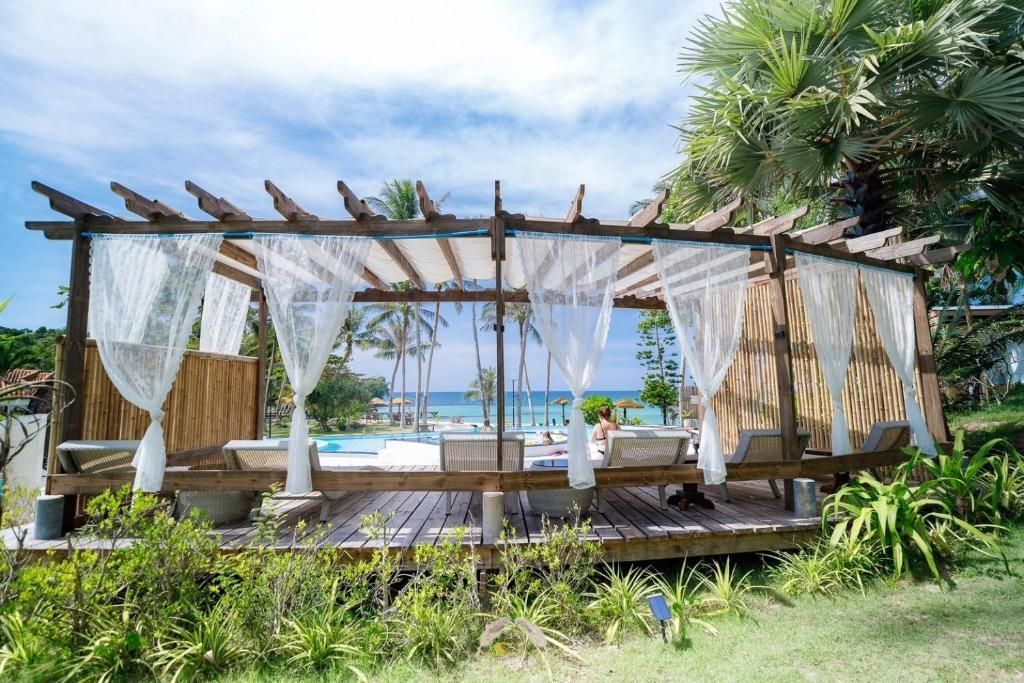 Wooden pergola with white curtains, overlooking a pool and ocean on a sunny day.