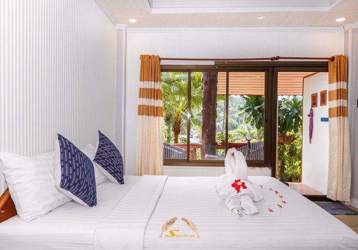 Bedroom with white bedding, pillows, and a towel swan, with a view of greenery.