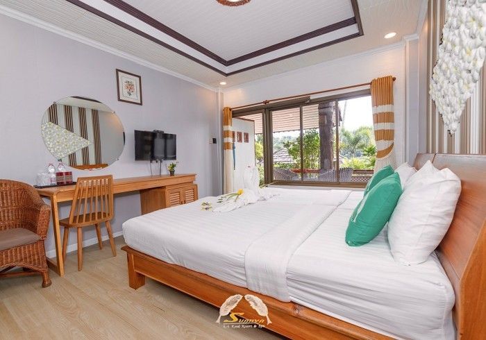 Bedroom with a large bed, desk, wicker chair, mirror, TV, and window with view.