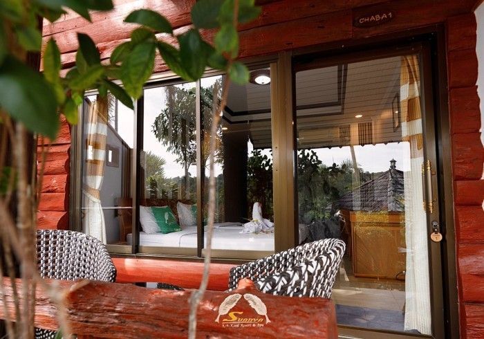 A cabin interior with a view of a bed and seating area. The exterior has red log siding and greenery.