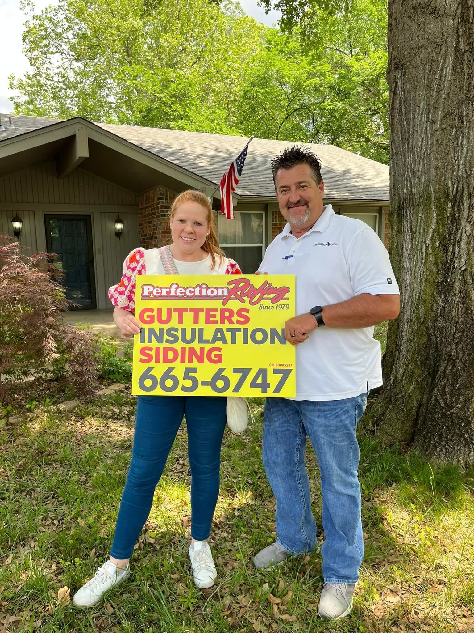 A man and a woman are standing in front of a house holding a sign.