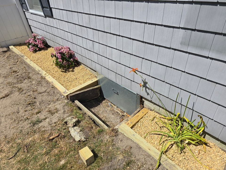 A house with a window well and flowers in front of it.