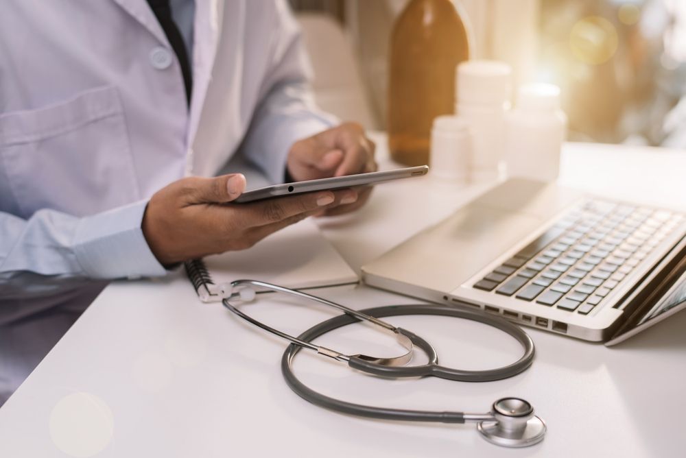 A doctor is using a tablet computer while sitting at a desk with a laptop and a stethoscope.