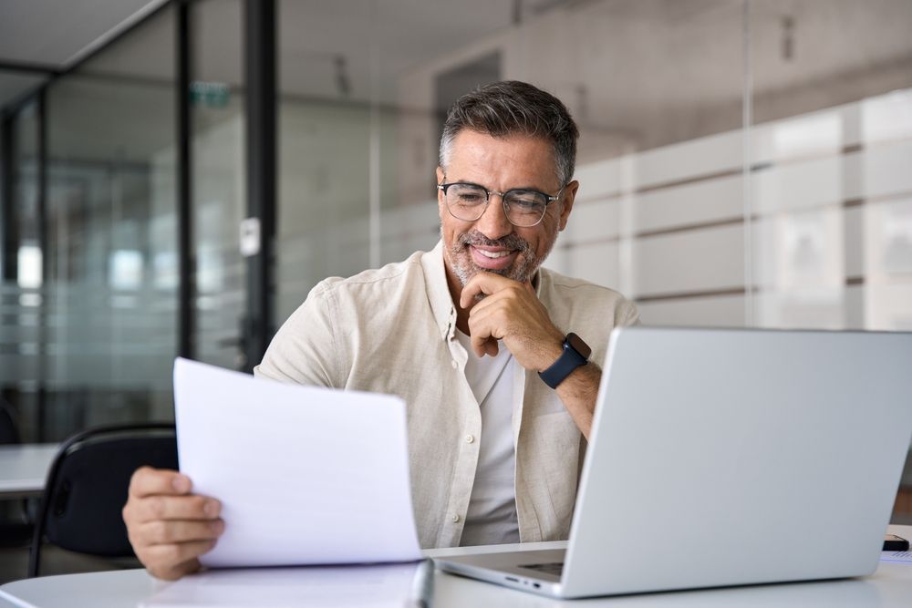 A man is sitting at a desk looking at a laptop and holding a piece of paper.