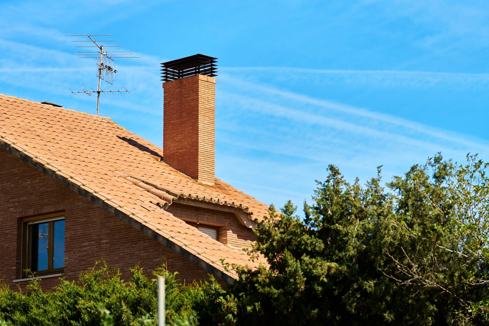 Brick house with red-tile roof, chimney, and antenna against a blue sky with contrails, trees in foreground.