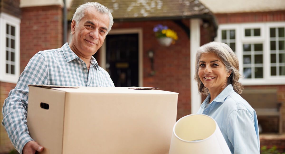 A man and a woman are standing in front of a house holding boxes.