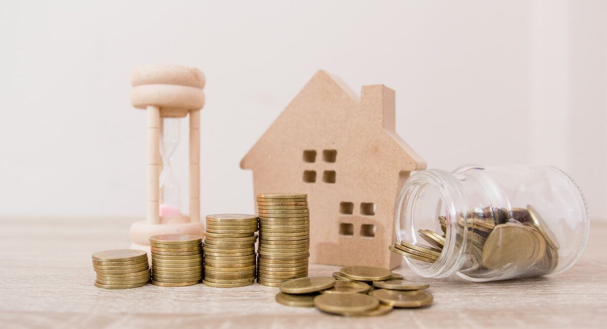 A wooden house , hourglass , stacks of coins and a jar of coins on a table.