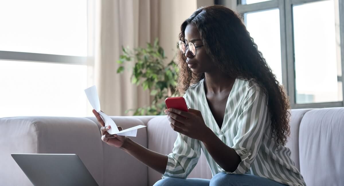 A woman is sitting on a couch using a laptop and a cell phone.