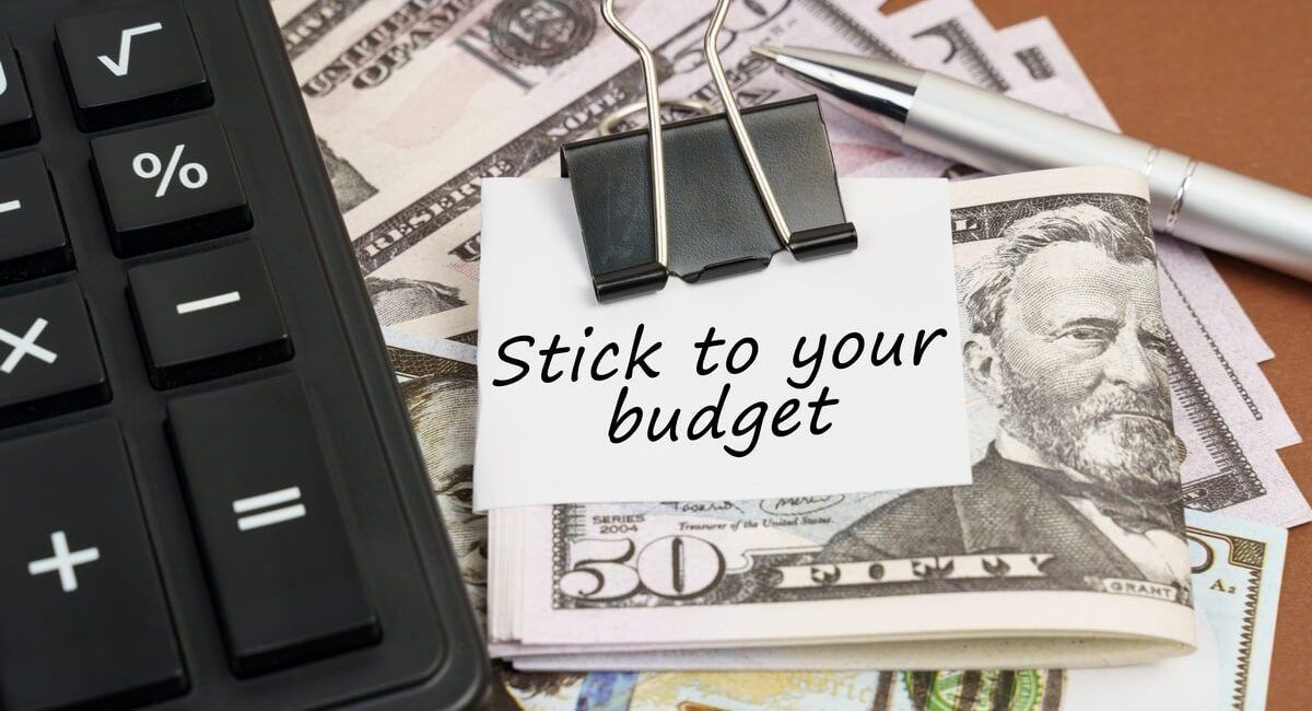 A note that says stick to your budget is sitting on top of a pile of money next to a calculator.