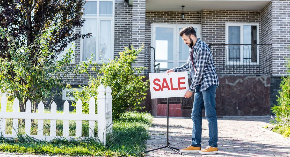 A man is standing in front of a house holding a sale sign.