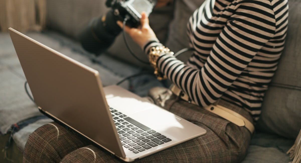 A woman is sitting on a couch using a laptop computer and holding a camera.