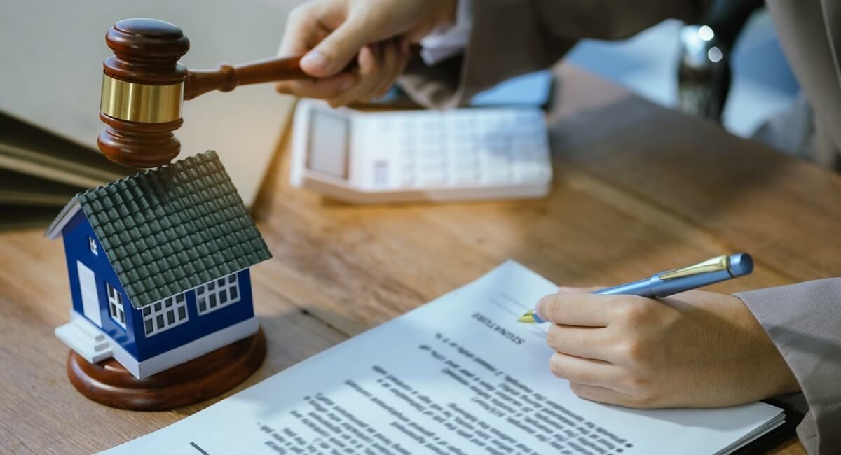 A person is writing on a piece of paper next to a model house and a judge 's gavel.