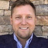 A man in a suit and blue shirt is smiling in front of a brick wall.