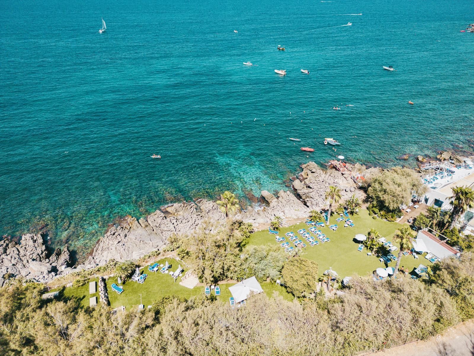 Vista aerea della costa rocciosa con prato verde, acqua turchese e barche.