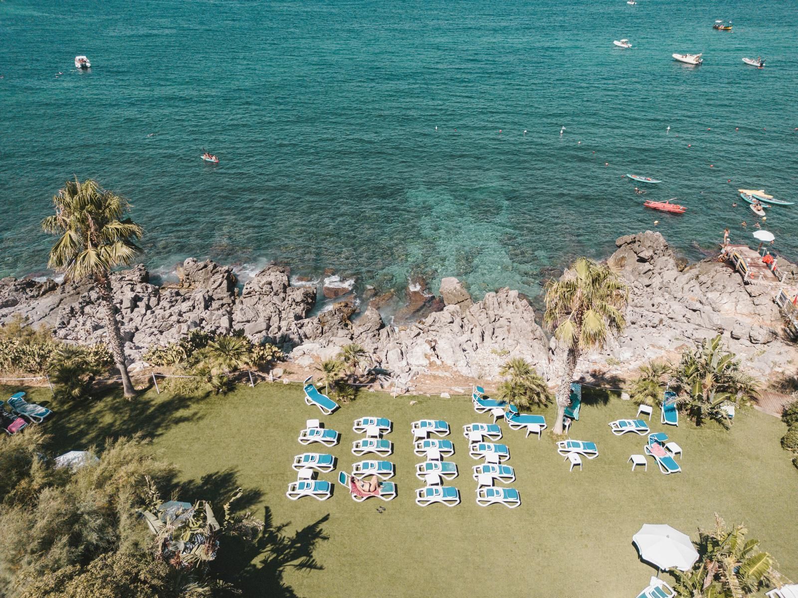 Un rigoglioso prato verde con sedie a sdraio vicino alla costa rocciosa e all'oceano turchese. Palme e barche sparse nell'acqua.