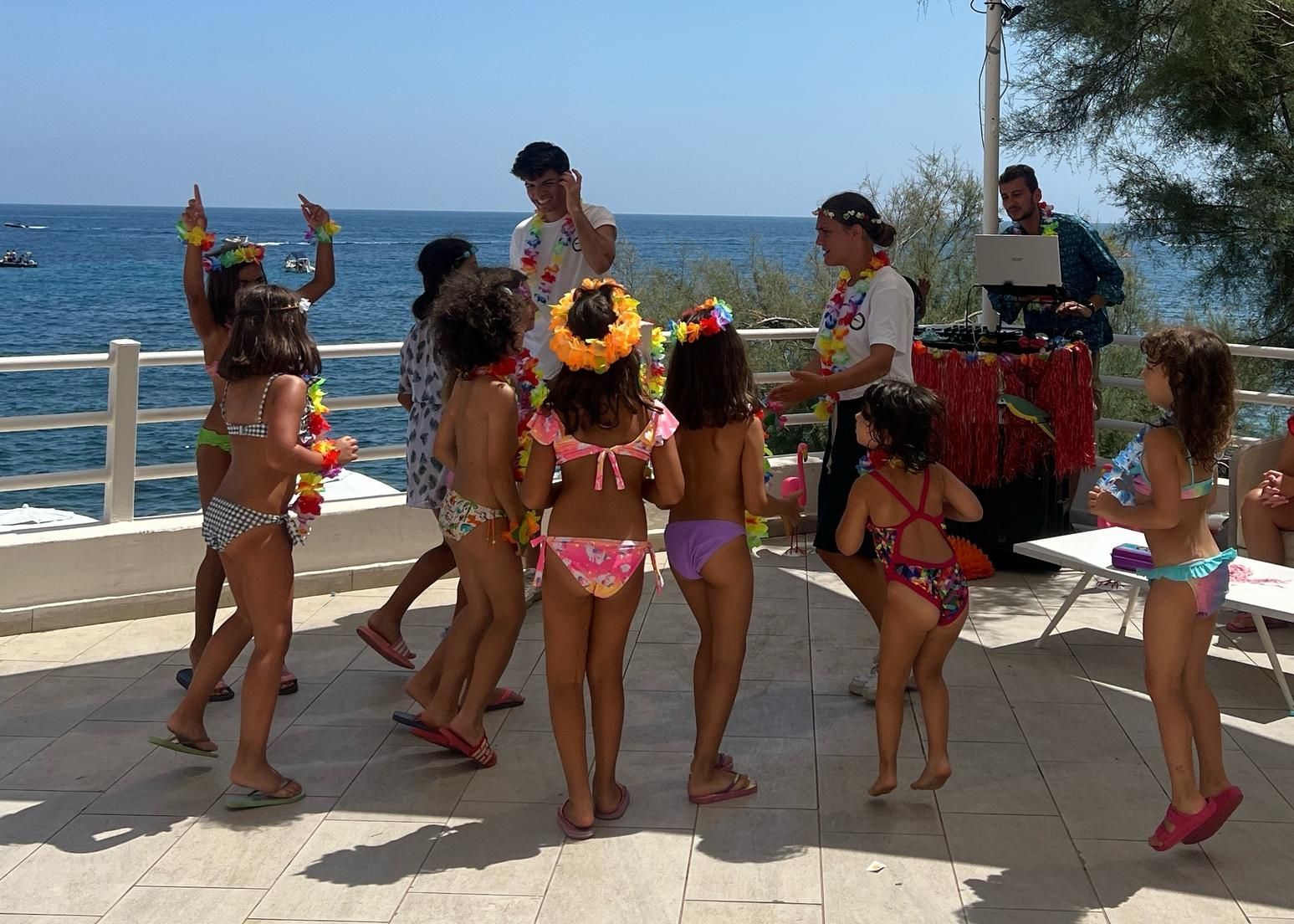 Bambini in costume da bagno che ballano all'aperto con gli adulti vicino all'oceano; sono presenti decorazioni festive.