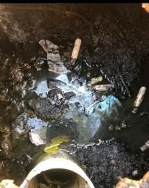 A view inside a dark, open drain containing standing water, debris, trash, and a white PVC pipe opening.