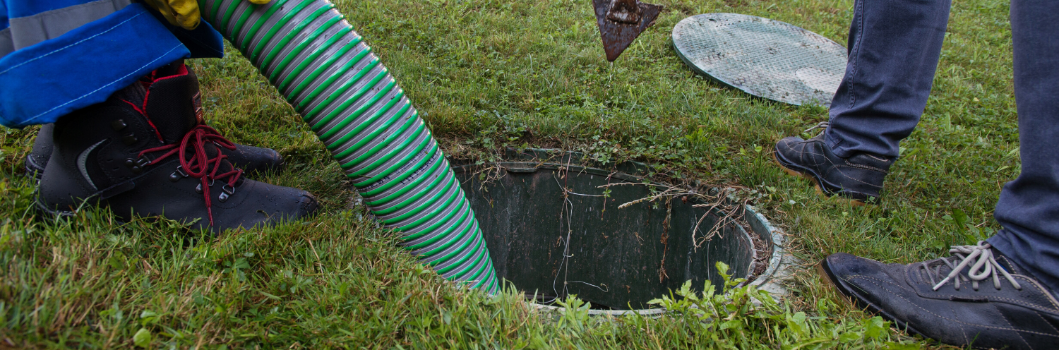 A worker uses a green vacuum hose to clean out an open residential septic tank in a grassy yard.