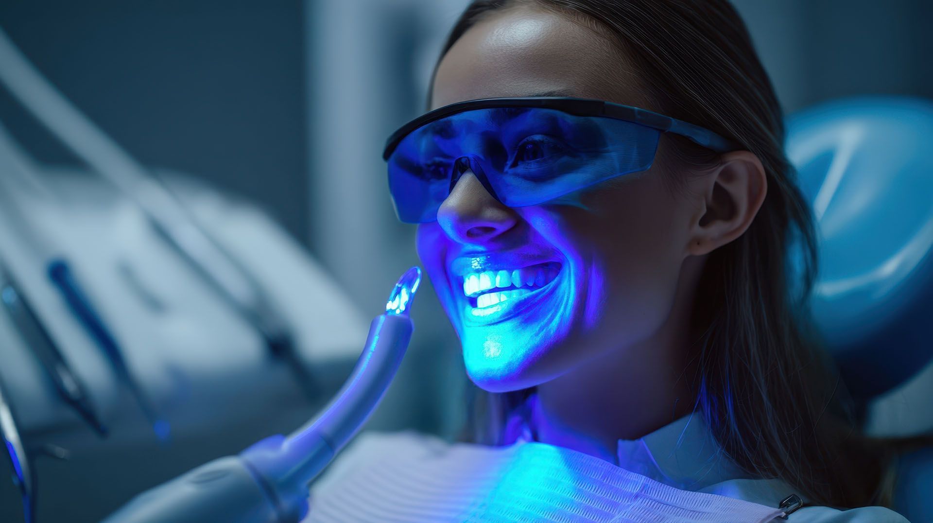 A smiling woman is receiving professional teeth whitening treatment with UV light and blue goggles.
