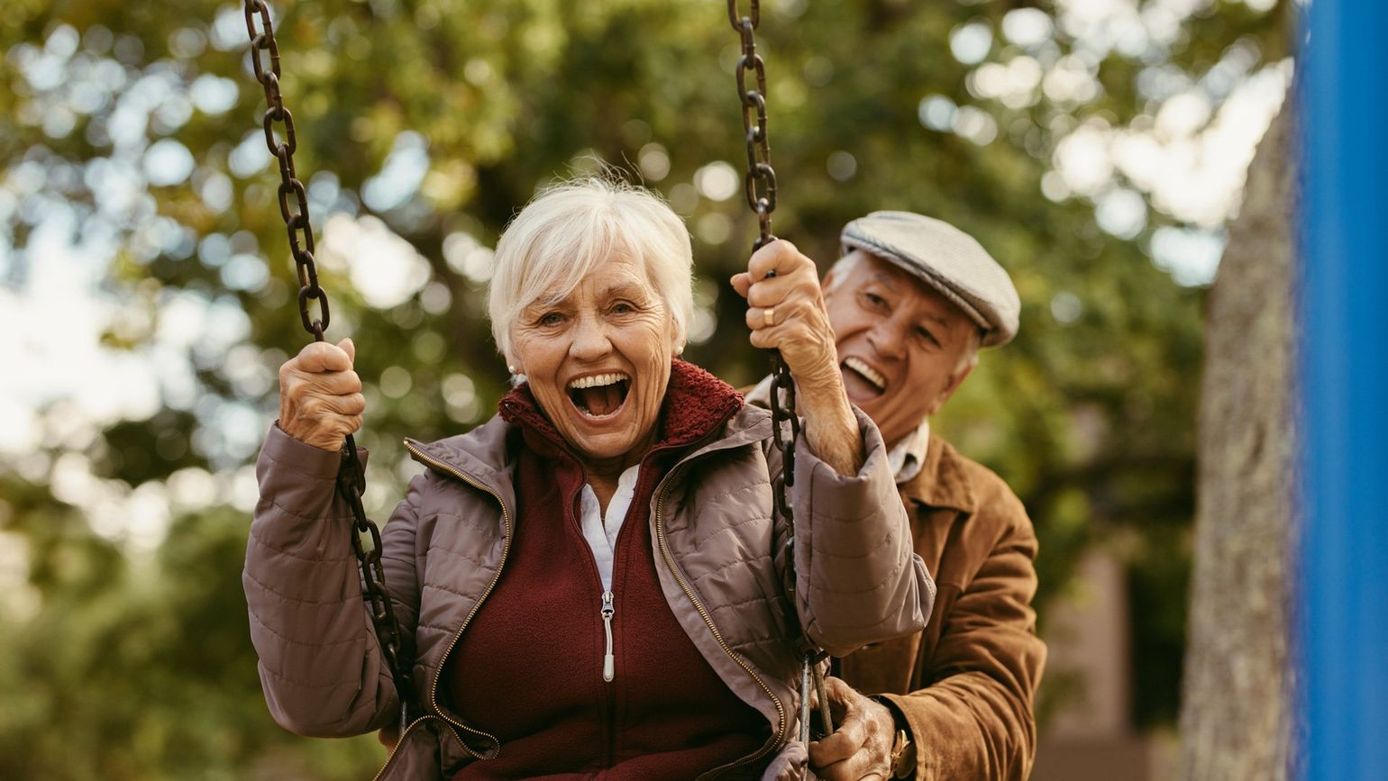 Elderly couple on a swing set, laughing with joy. Woman is swinging, man is pushing. Outdoors, green background.