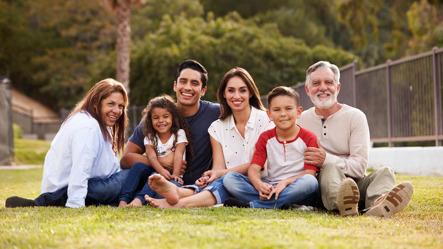 Family of seven smiles for the camera, sitting on grass in a park.