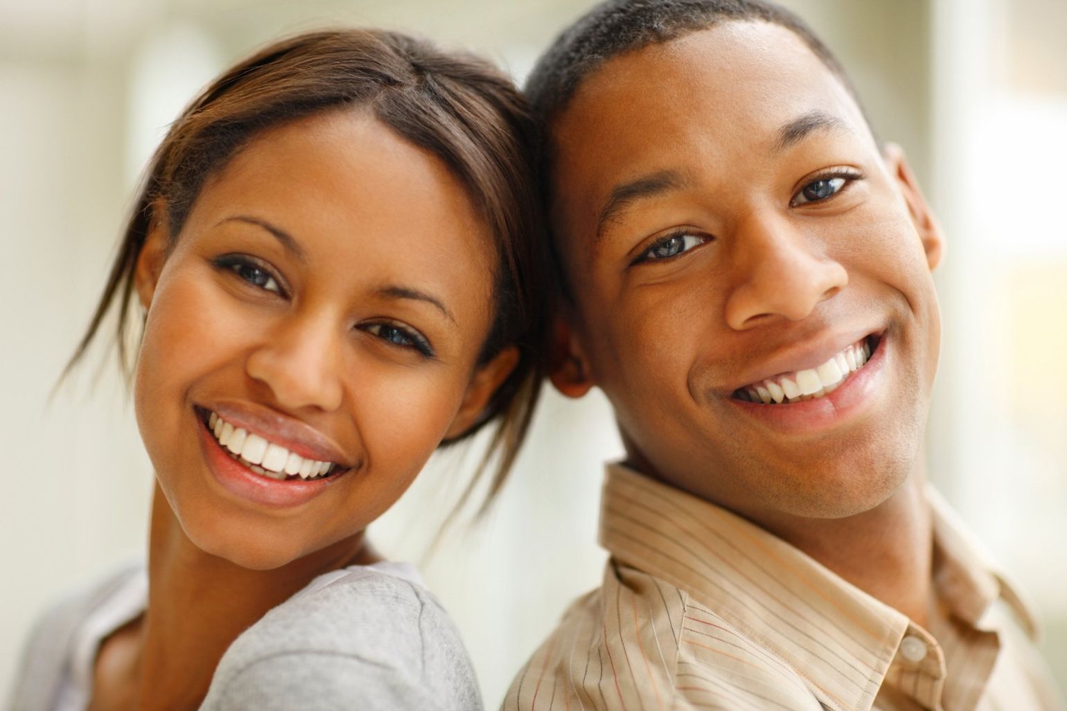 Smiling couple, backs touching, looking at the viewer. One wearing a beige shirt, the other a gray top.