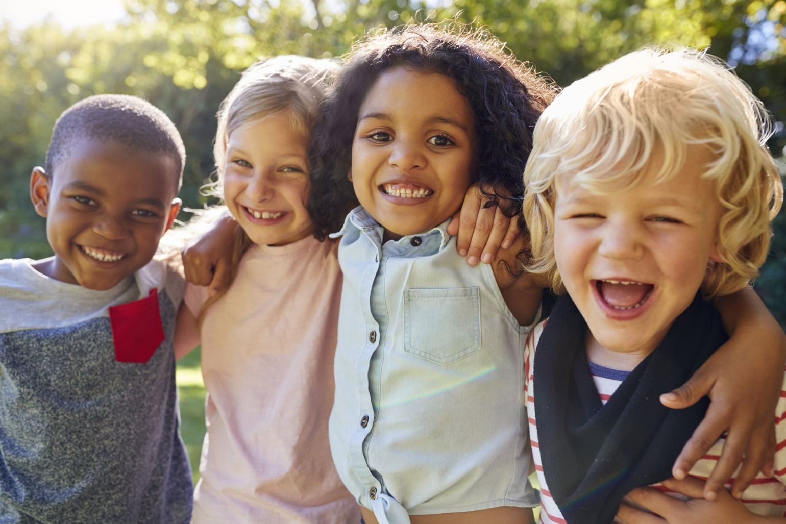 Four smiling children with arms around each other, outdoors.
