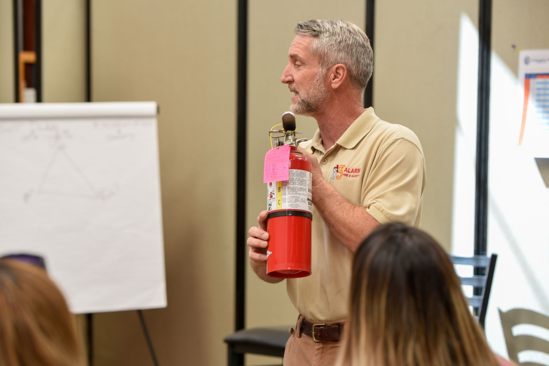 A person in a tan polo shirt holds and demonstrates a red fire extinguisher in front of an audience.