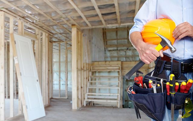 Construction worker holding a hammer and hard hat in a partially built house; wooden framing and a door are visible.
