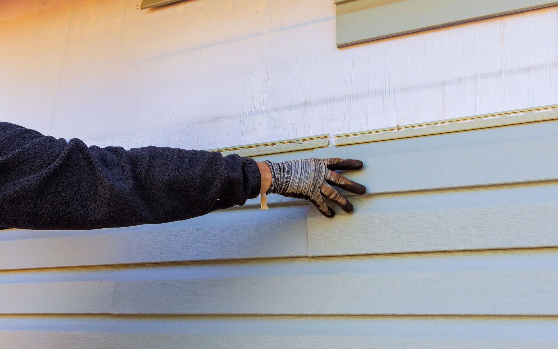 Person's gloved hand installing light blue siding on a house.