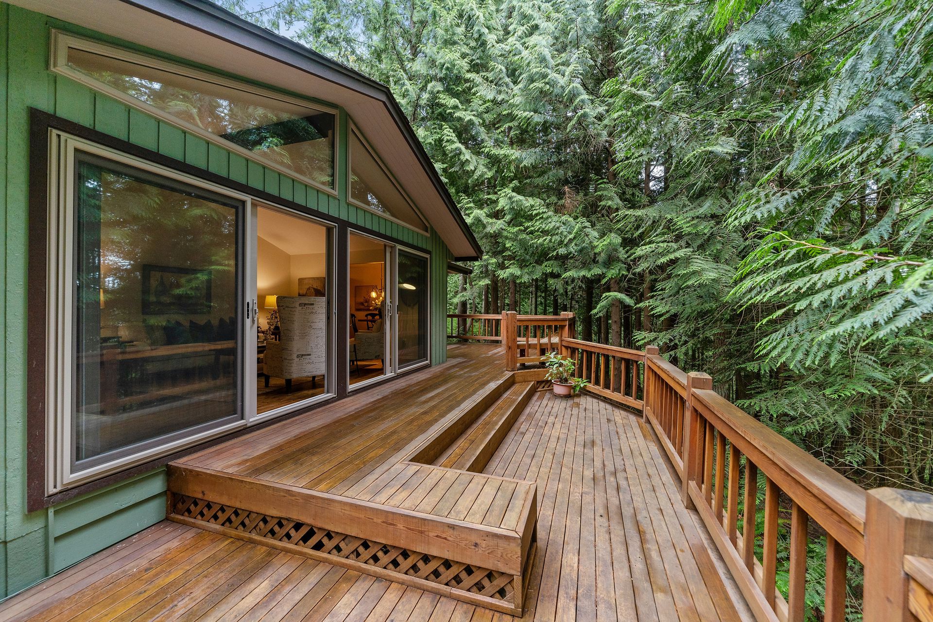 A green house with a large wooden deck in front of it surrounded by trees.