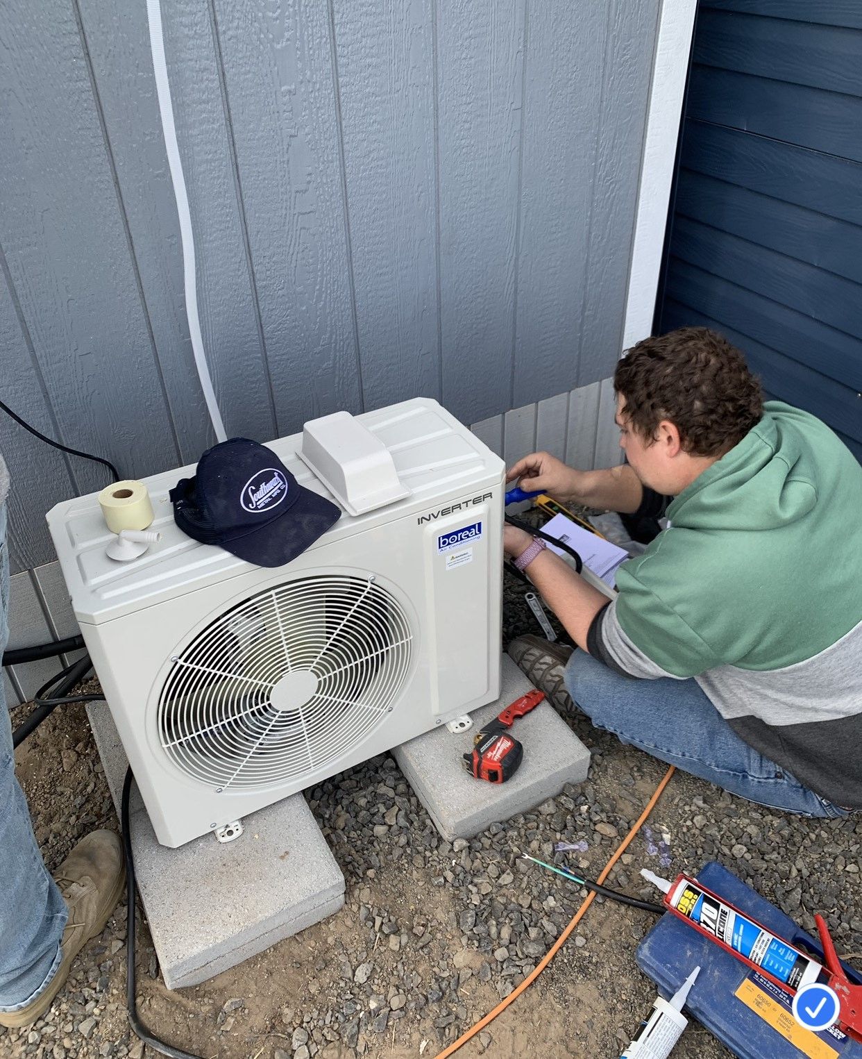 HVAC technician kneels beside AC unit, working outdoors. He wears gloves, a hat, and work overalls.