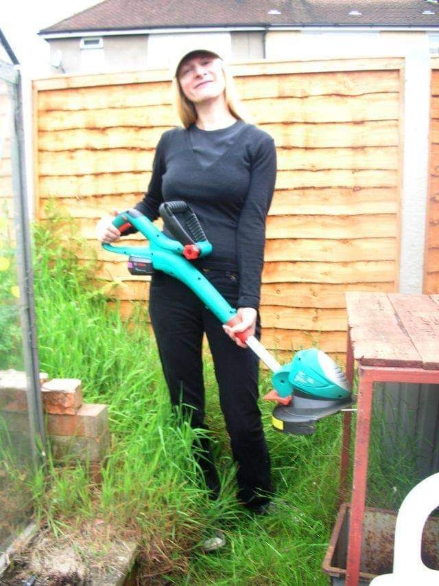 Woman dressed in black jeans and top, baseball cap on top of head, holding a garden strimmer, standing in an overgrown garden.