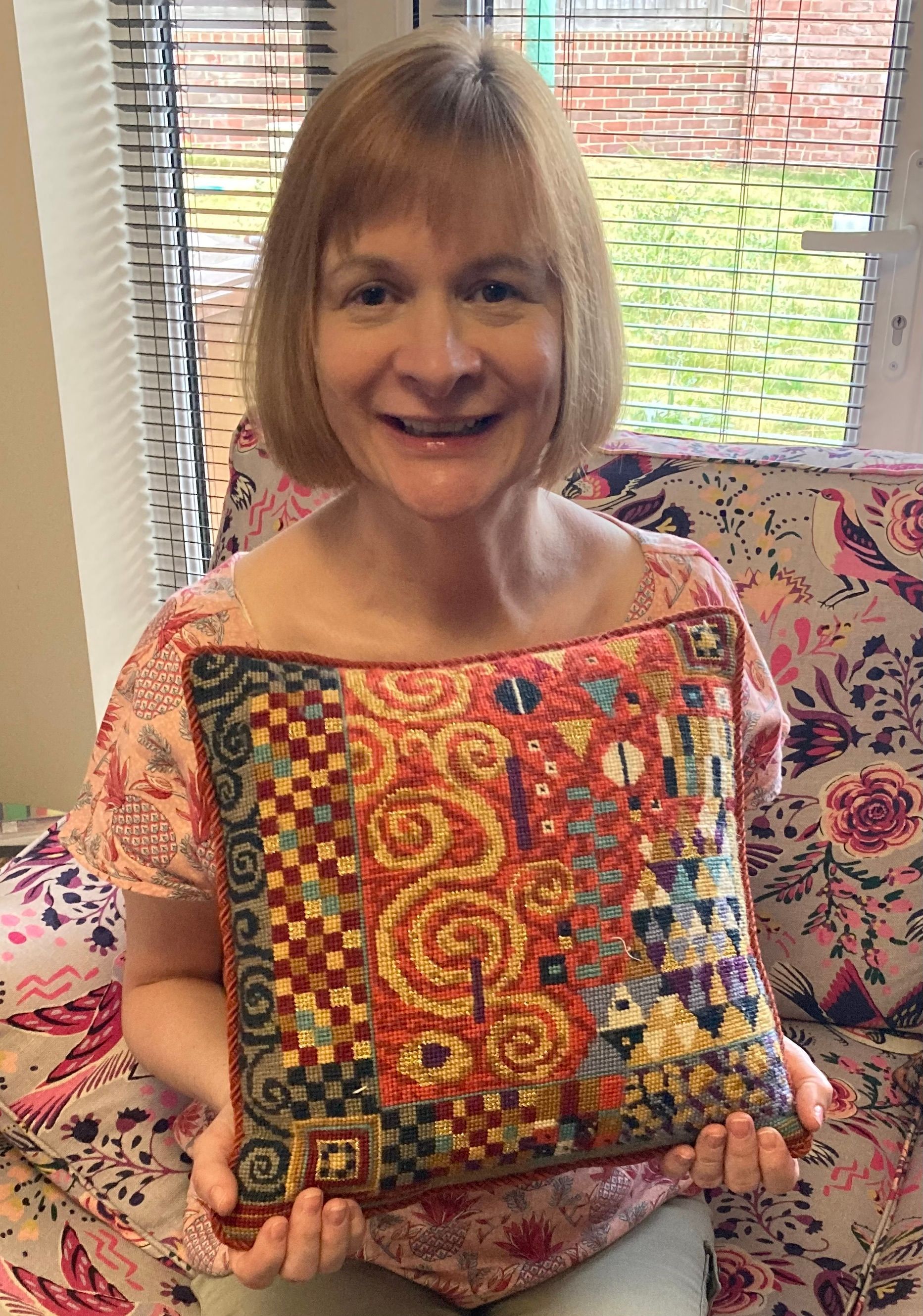Woman sitting on a sofa holding a cross-stitched, handmade, colourful cushion. Cushion design based on the work of Gustav Klimt.