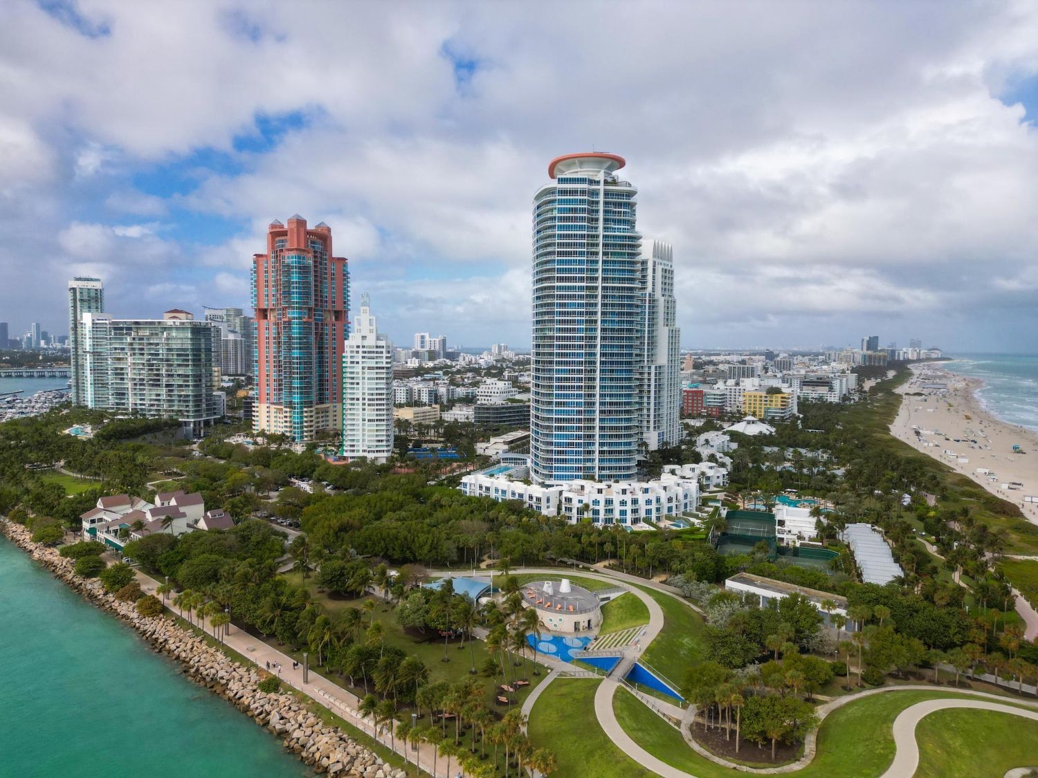 Miami Beach skyline with tall buildings, beach, ocean, and green park under a cloudy sky.