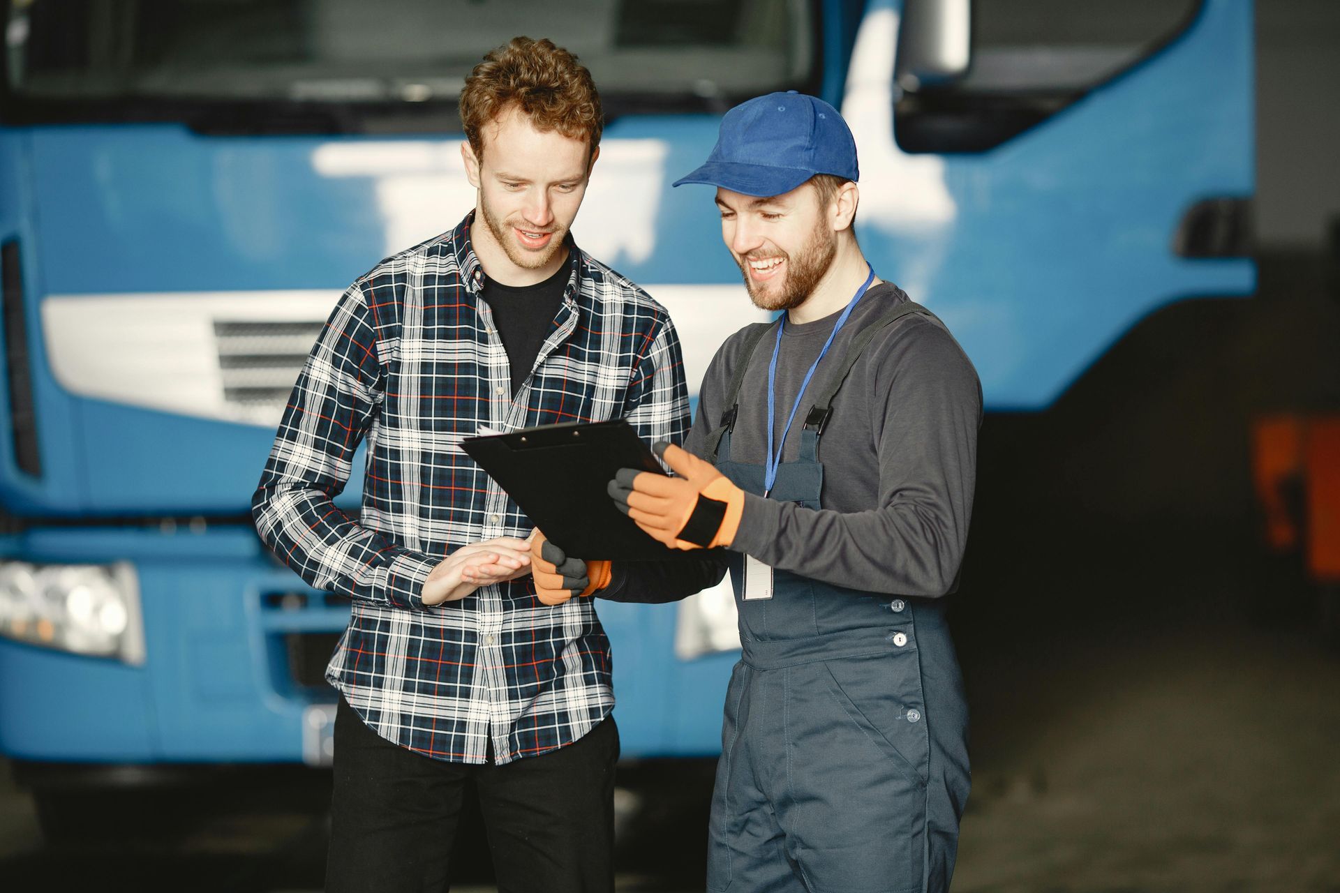 Two men looking at a clipboard in front of a blue truck. One man wears a plaid shirt, the other overalls and a blue cap.