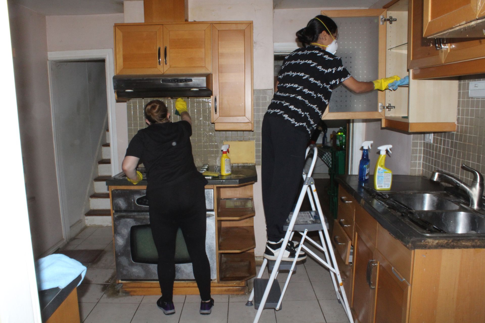 Two people cleaning a kitchen: one on a ladder wiping cabinets, the other cleaning the stove.
