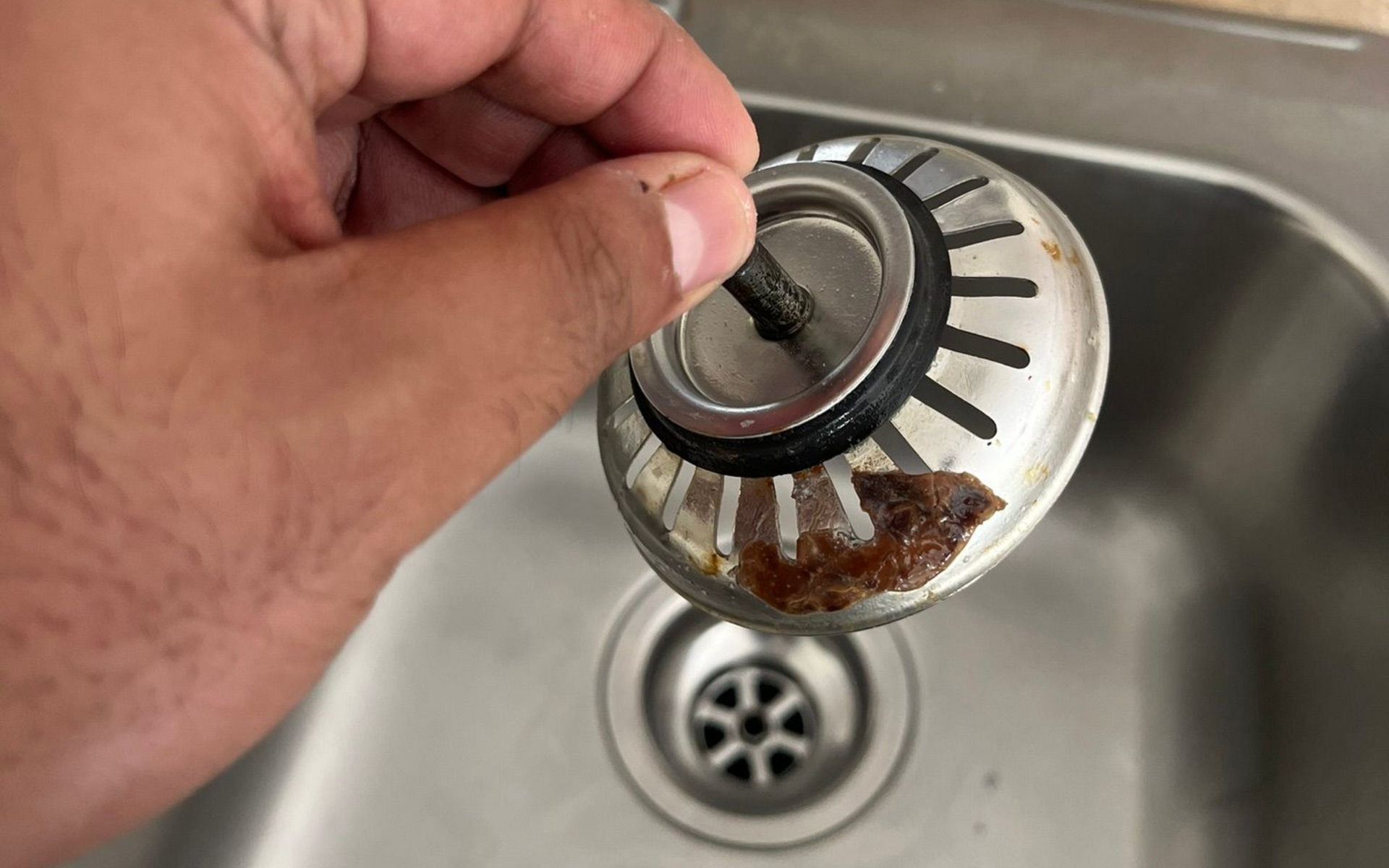 Hand holding a dirty sink strainer with debris over a stainless steel sink.