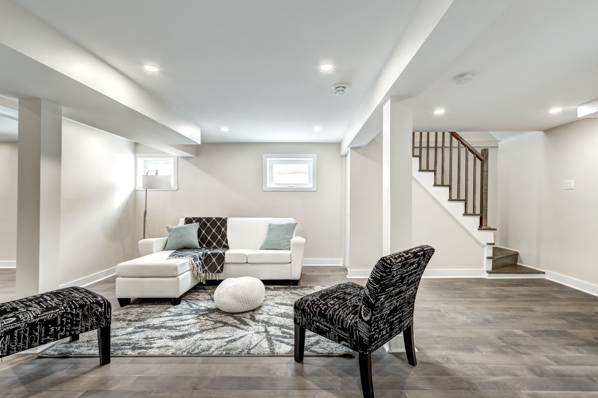 Bright modern living room with white sofa, patterned chairs, gray rug, and staircase in the background