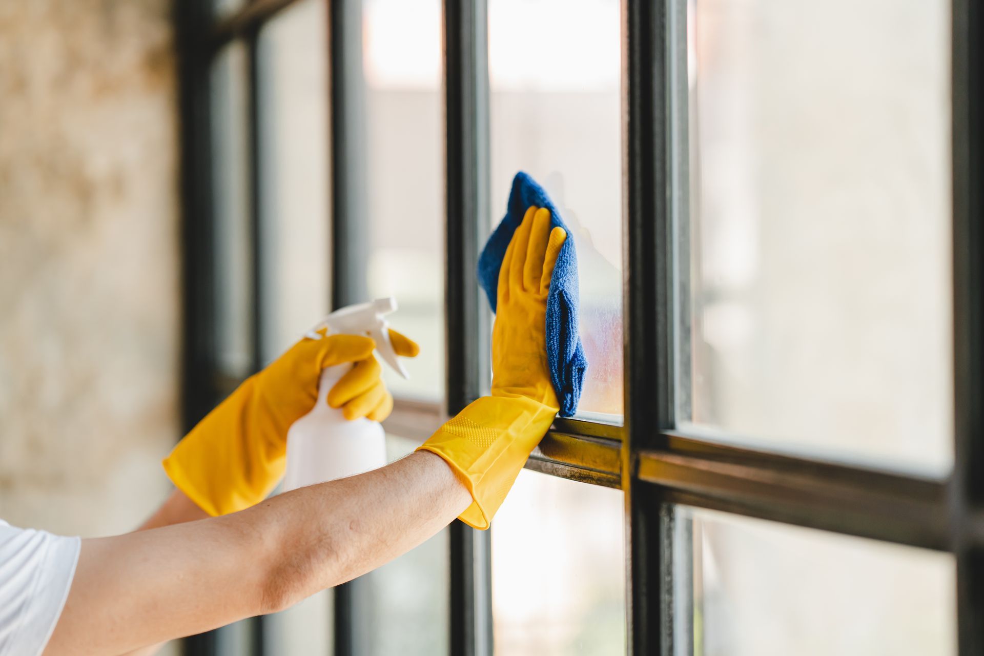 Person wearing yellow gloves spraying and wiping a window with a blue cloth.