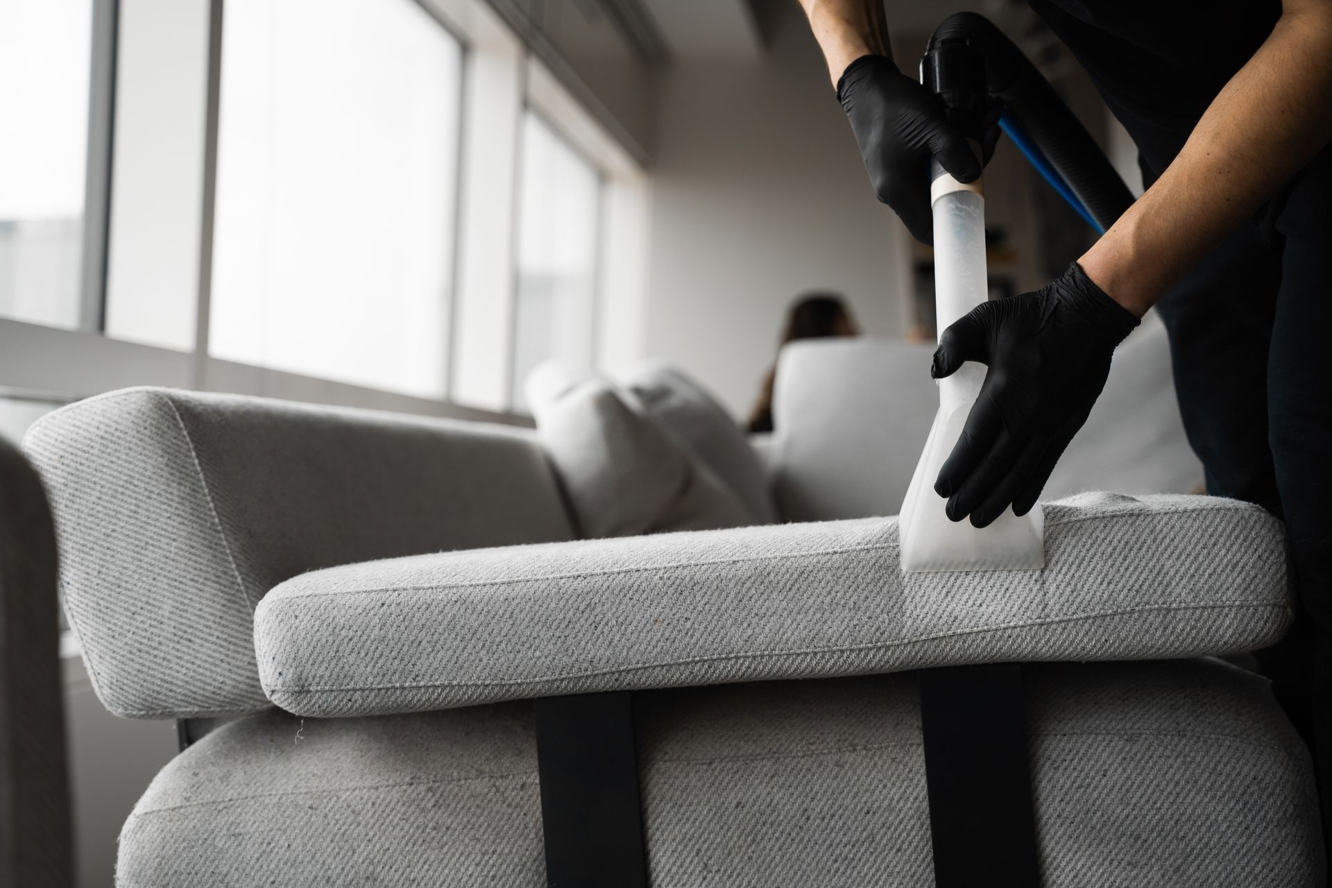 Person in black gloves cleaning a gray sofa cushion with a cleaning tool indoors.