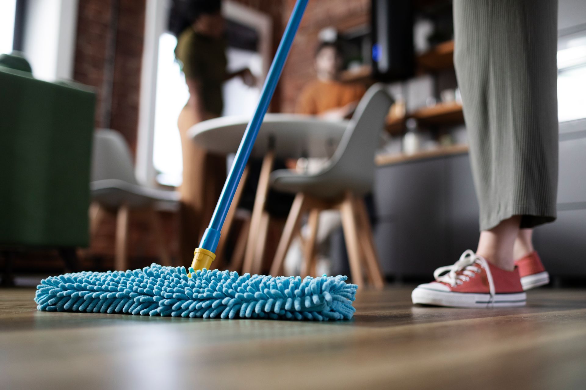 Person mopping a wooden floor, blue mop in focus. Two people in the blurred background.