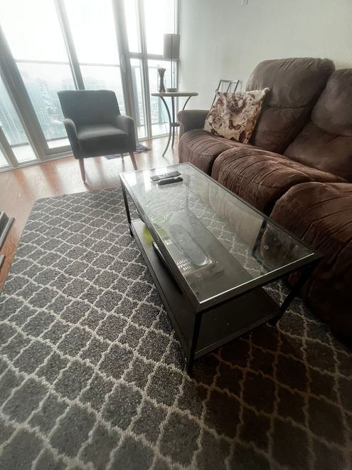 Living room: brown recliner sofa, gray patterned rug, glass coffee table, armchair, and a city view.