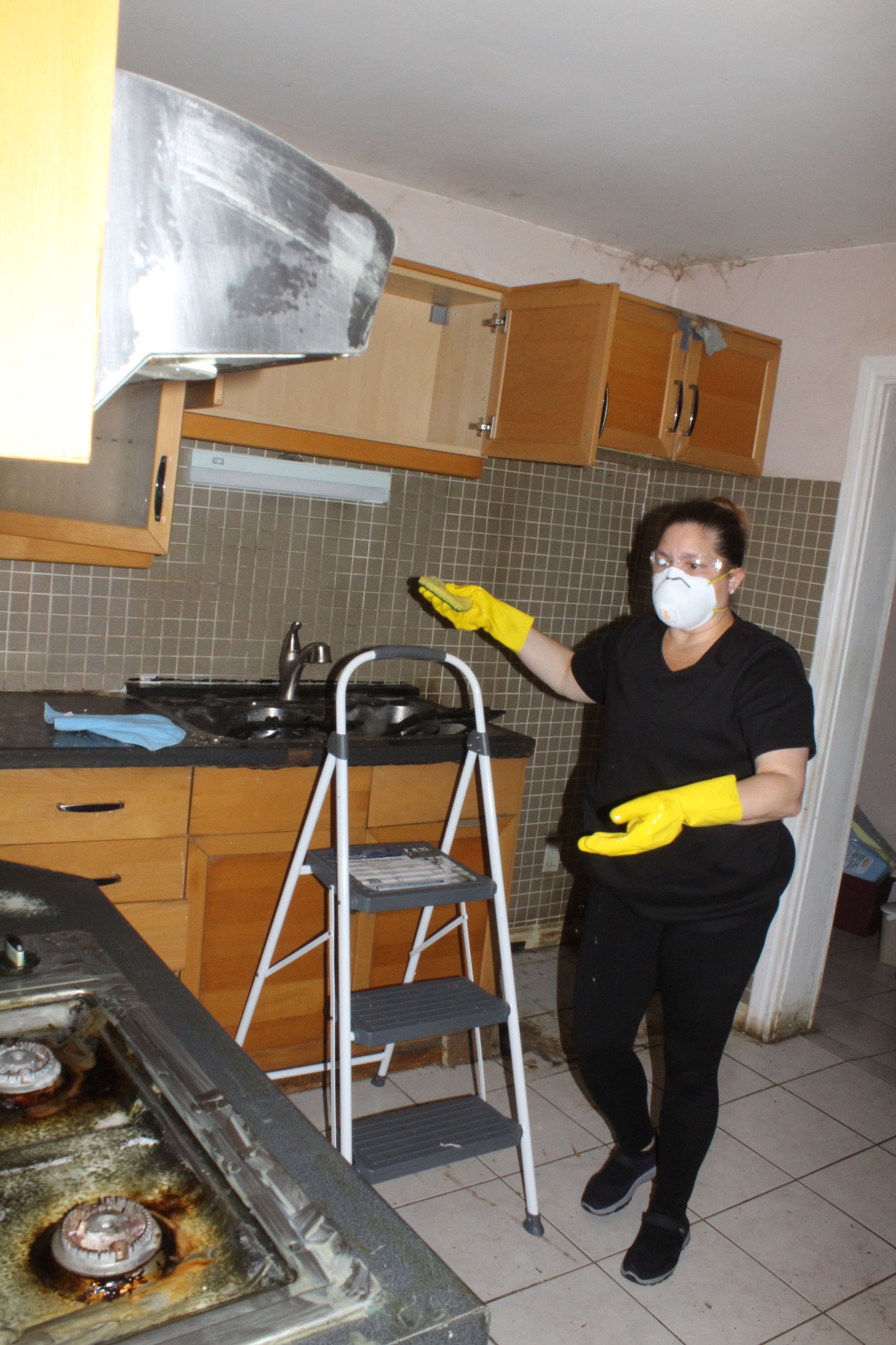 Woman in mask and gloves pointing at damaged kitchen cabinets, next to a step ladder and stove.